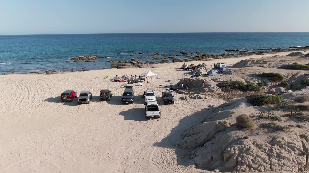 At 0:02, vehicles remain grouped on the sand while people cluster near the white canopy, with low tan rocks in the foreground and blue water across the background.