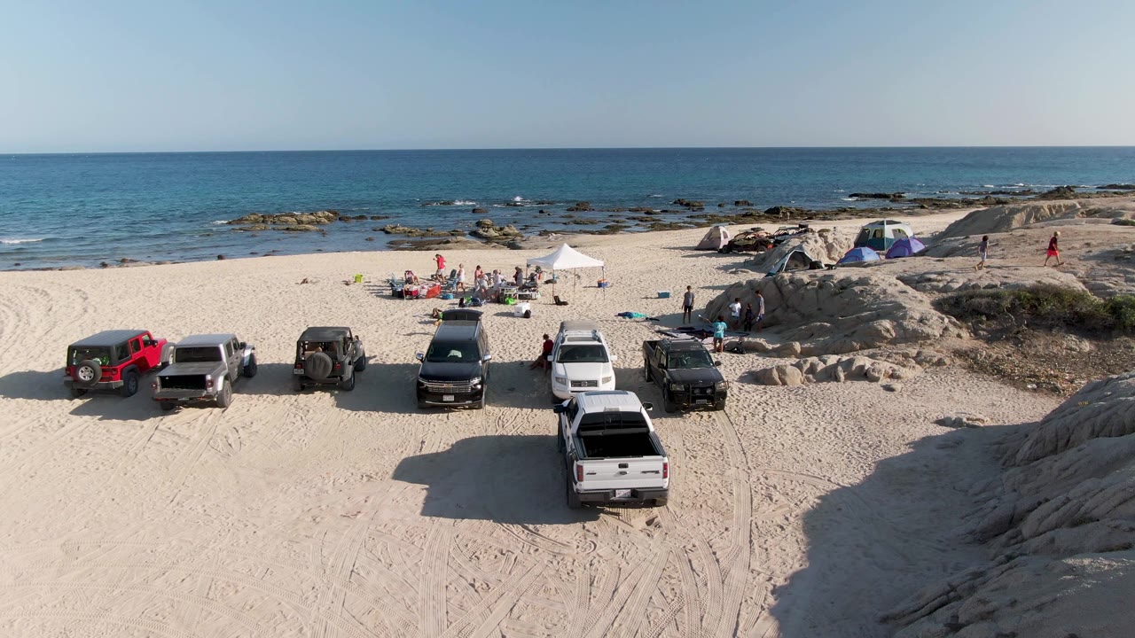 At Shipwrecks Beach in Mexico, an elevated view shows six parked vehicles on sand, small groups near tents and shade canopies, rocky shoreline, and blue ocean behind them.