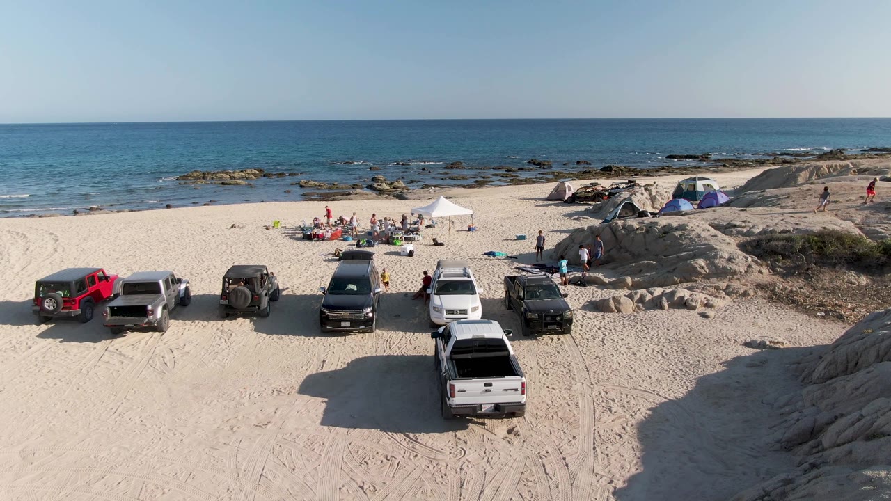 At 0:02, parked vehicles remain lined across the sandy foreground while beachgoers cluster around tents and a white shade canopy near the rocky waterline at Shipwrecks Beach.
