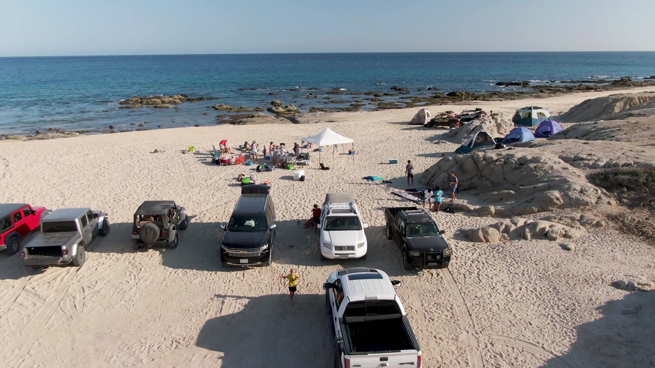 At Shipwrecks Beach on the East Cape of Los Cabos, Mexico, an elevated view shows parked trucks on sand, a white canopy with people, tents near dunes, rocky shore, and blue ocean.