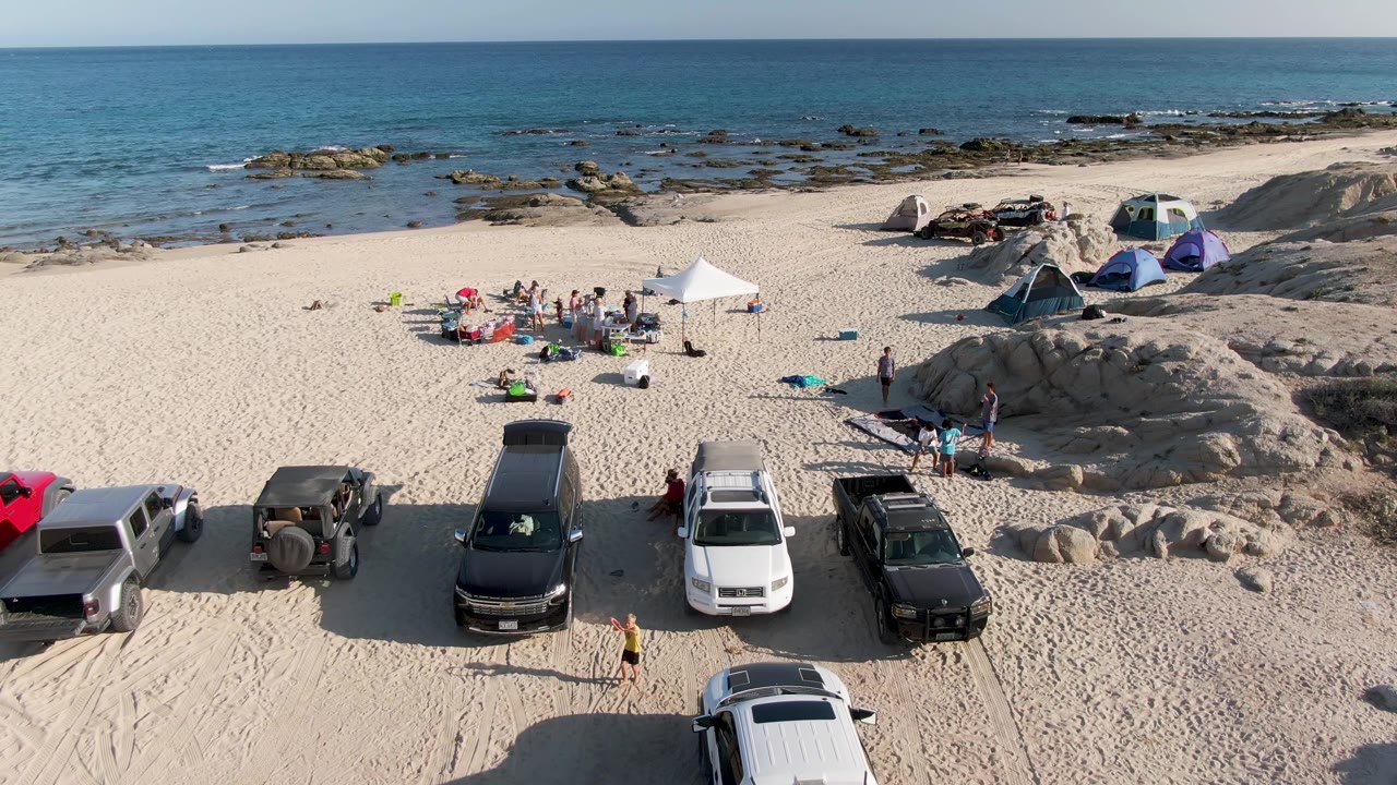 At 0:02, the aerial composition holds on Shipwrecks Beach with trucks and SUVs on the sand, small figures near a canopy, tents along the dune edge, scattered rocks, and calm blue water.