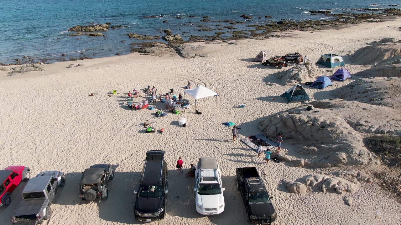 At Shipwrecks Beach East Cape Los Cabos BCS Mexico, an overhead frame shows people gathered near a white canopy, tents to the right, parked vehicles below, and rocky blue water above.