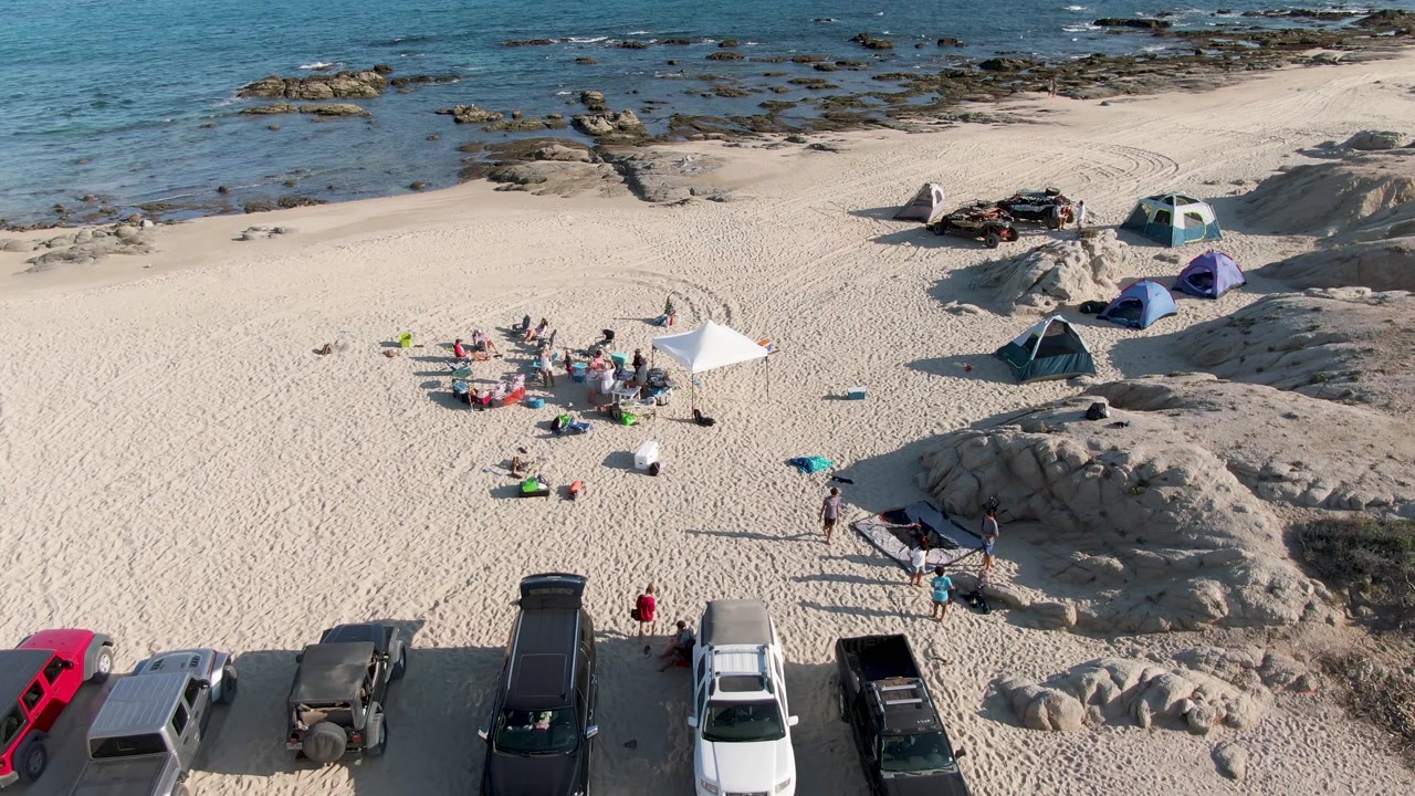 Overhead beach scene with people near a canopy and parked cars along the sand.