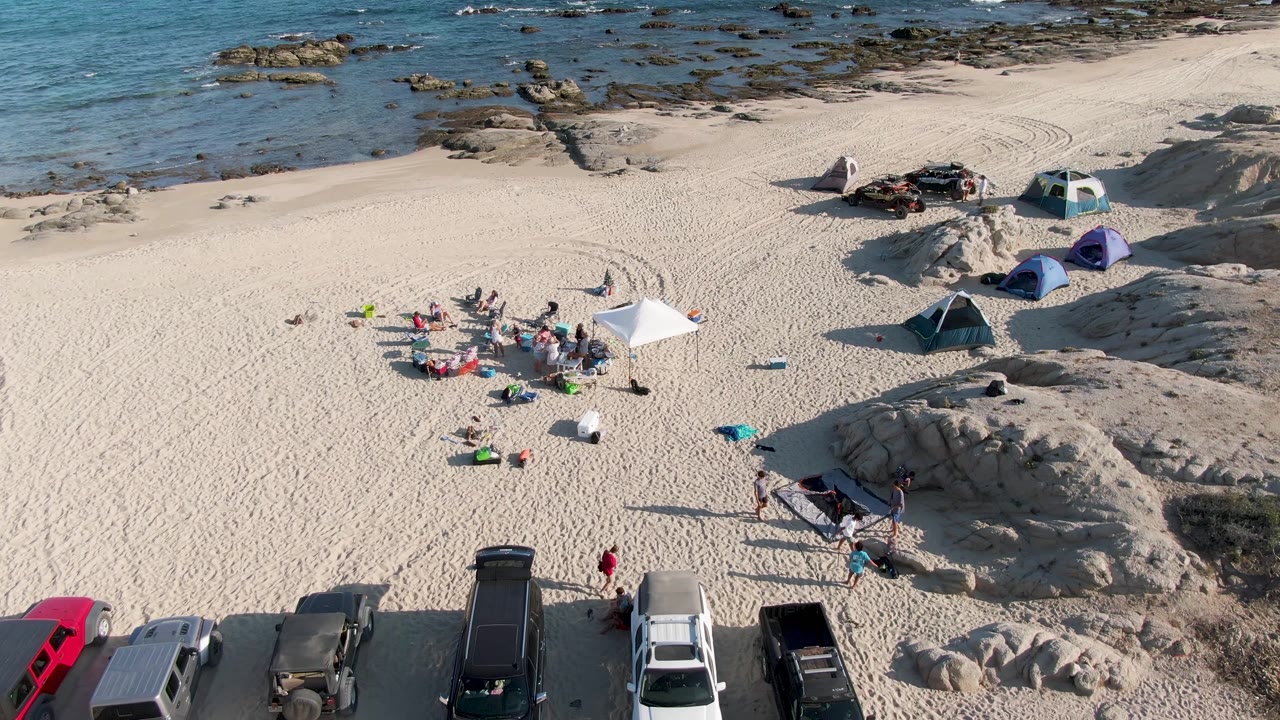 At Shipwrecks Beach, the 0:02 frame keeps the shoreline rocks high in frame while the beach group, white canopy, tents, and a row of parked vehicles remain visible below.