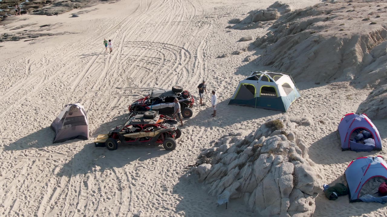 Aerial frame at Shipwrecks Beach in Mexico showing multiple tents, parked ATVs, visible people, tire-marked sand, pale rock formations, and ocean surf at the top edge.