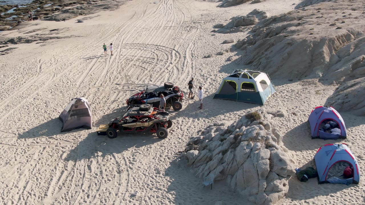 Overhead beach campsite with tents, ATVs, walking people, sand tracks, and rocky terrain.