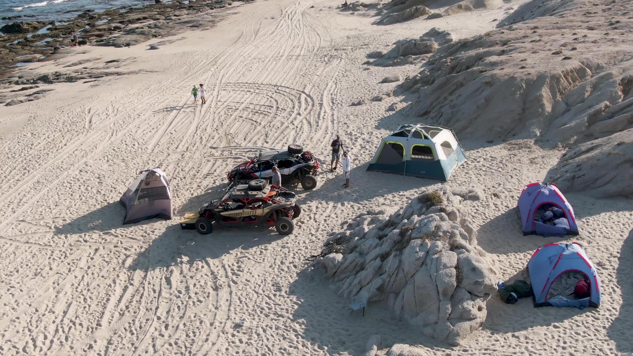 Aerial view at Shipwrecks Beach in Mexico showing the campsite from a similar high angle, with pink and blue tents at right, gray tent at left, ATVs near center, people in the sand, and waves more visible along the top.