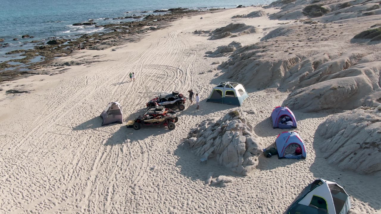 Aerial frame at Shipwrecks Beach East Cape Los Cabos showing several tents around a sandy campsite, small people near vehicles, rounded rocks, and the ocean beyond.