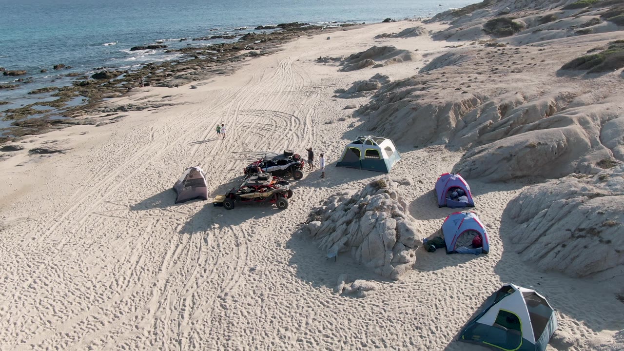 Beach campsite with tents, people, vehicle, sand tracks, rocks, and ocean in the background.