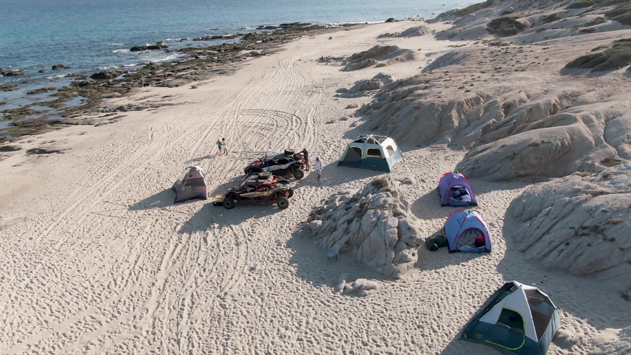 Aerial frame at Shipwrecks Beach East Cape Los Cabos BCS Mexico showing multiple tents, an all-terrain vehicle with gear, pale sand tracks, rocks, ocean water, and small visible people near the camp.