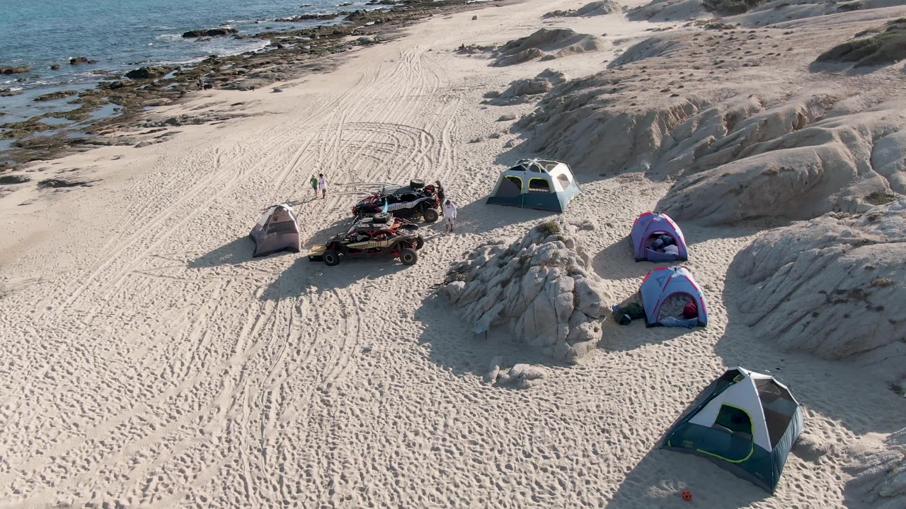 Overhead frame at Shipwrecks Beach East Cape Los Cabos BCS Mexico showing tents spaced around a loaded all-terrain vehicle, curving tire tracks in sand, low rock ridges, ocean water, and tiny visible people.