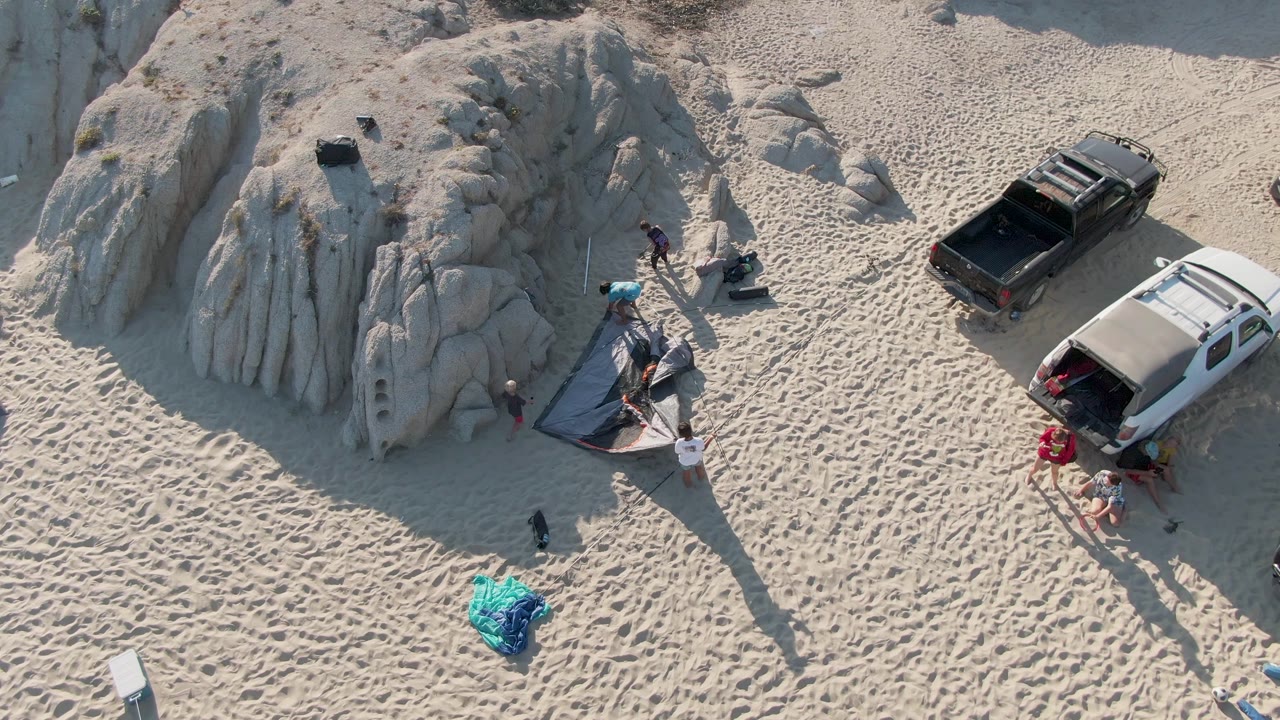 Overhead frame at Shipwrecks Beach East Cape Los Cabos BCS Mexico showing people around a dark tent beside a pale rock formation, with a black pickup, white van, blue bundle, and long shadows on sand.