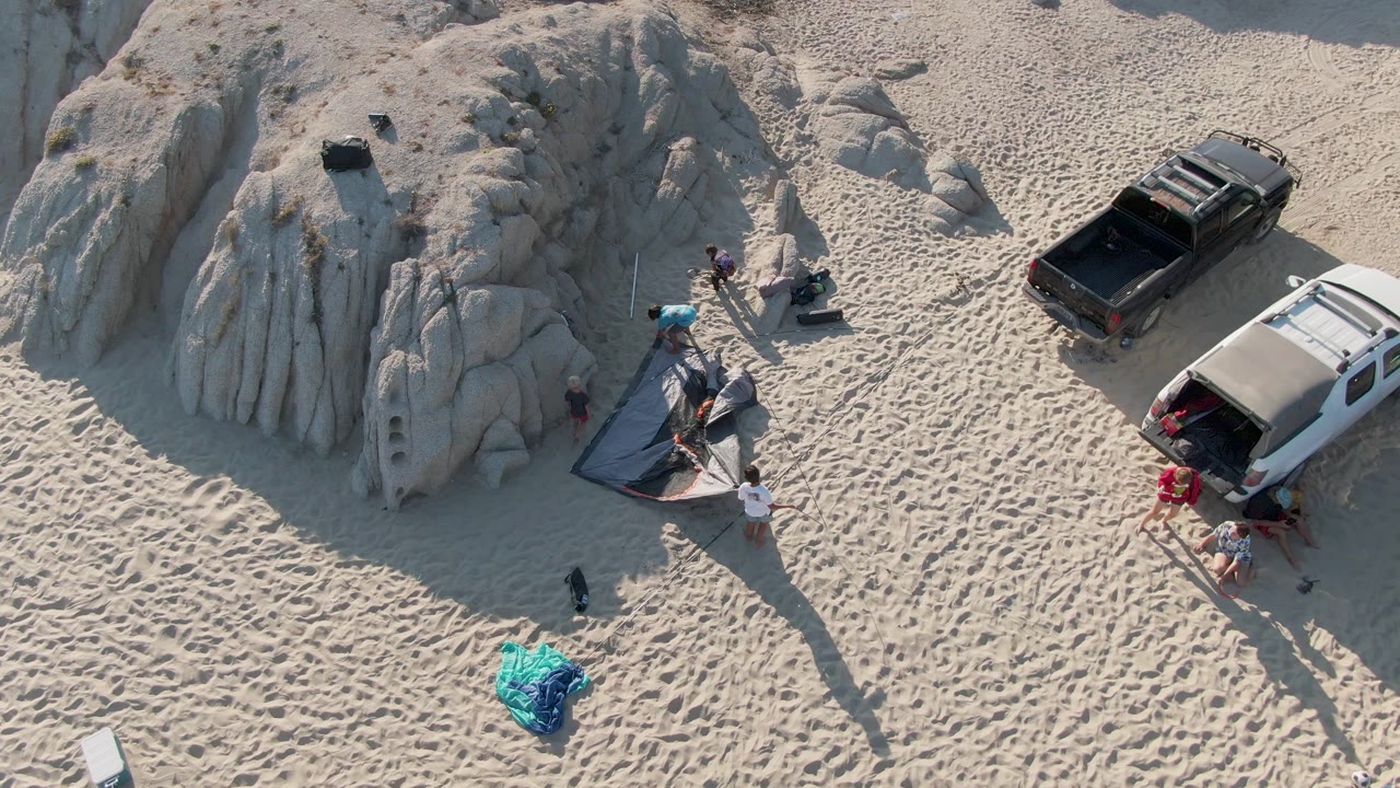 People stand and move near a tent on sand beside a rock formation and two parked vehicles.