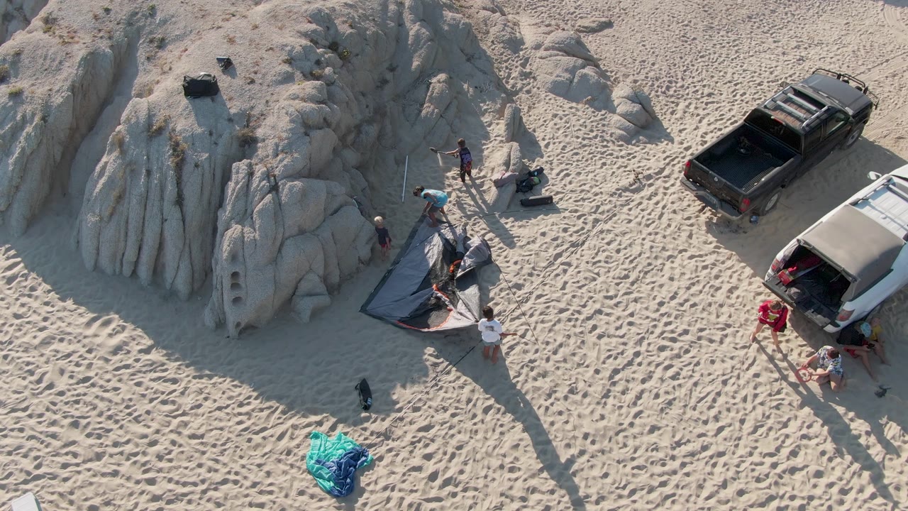 Overhead frame at Shipwrecks Beach East Cape Los Cabos BCS Mexico showing the group clustered near the tent, with the black pickup and white van separated by the rock formation and deep footprints in the sand.