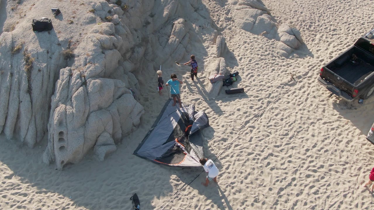 Overhead frame at Shipwrecks Beach in Mexico showing several people around a partly spread tent beside pale rock formations and textured sand.