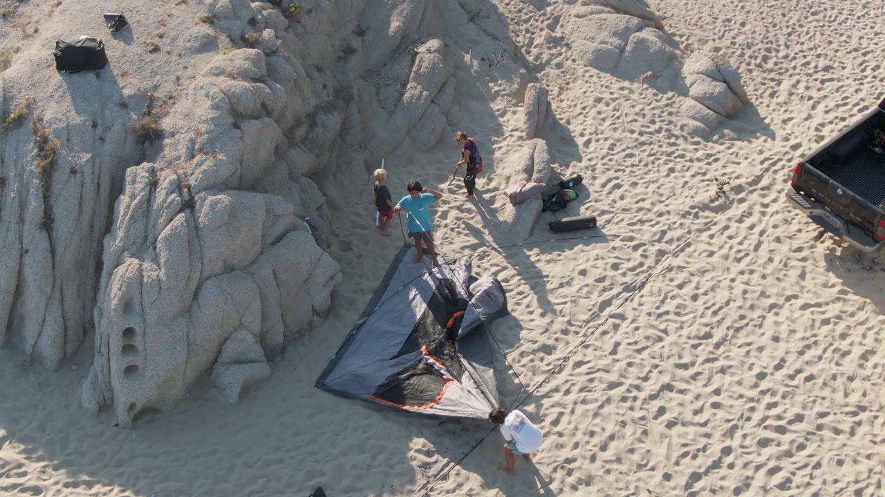 Beachgoers stand around tent fabric near rocks and gear.