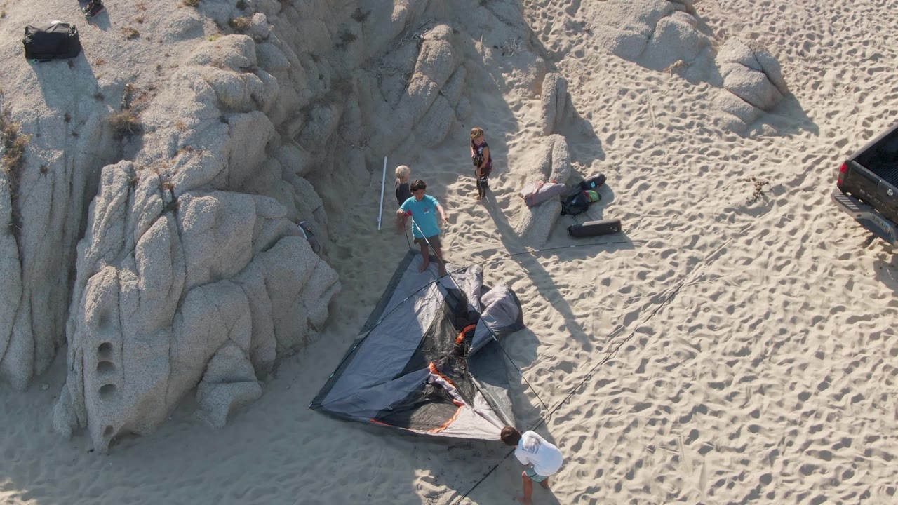 Shipwrecks Beach overhead frame showing the group continuing to work around the tent, with one person near the lower edge and others beside rocks and gear.
