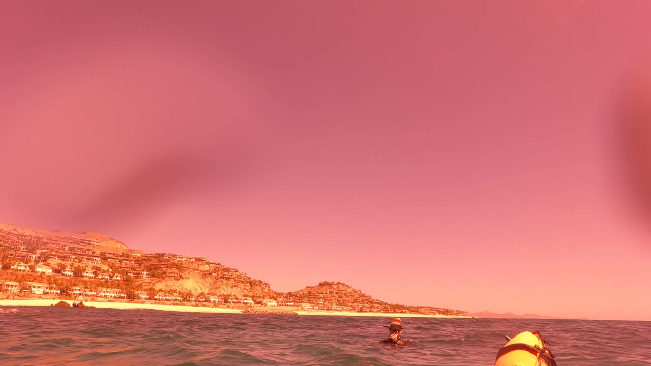 Water-level view in San Jose del Cabo BCS Mexico showing a snorkeler near the surface, small waves, warm pink light, and a hilly coastline behind the beach.