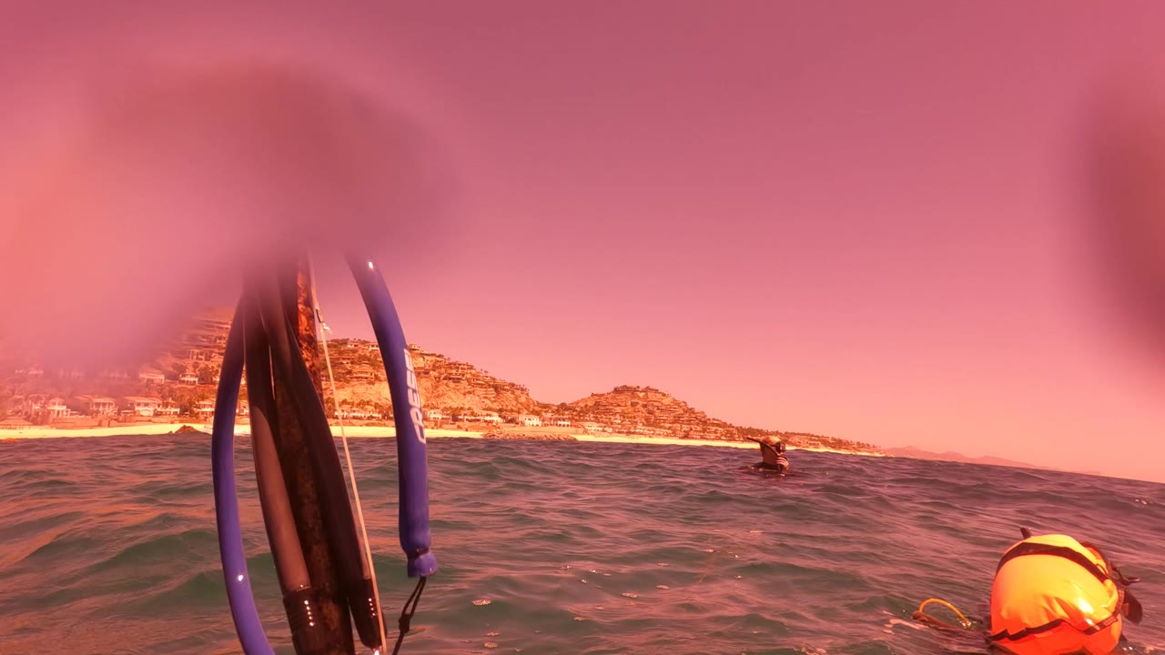 Foreground snorkel gear rises near the lens while a swimmer remains beyond the waves, with San Jose del Cabo BCS Mexico beach and hills across the horizon.
