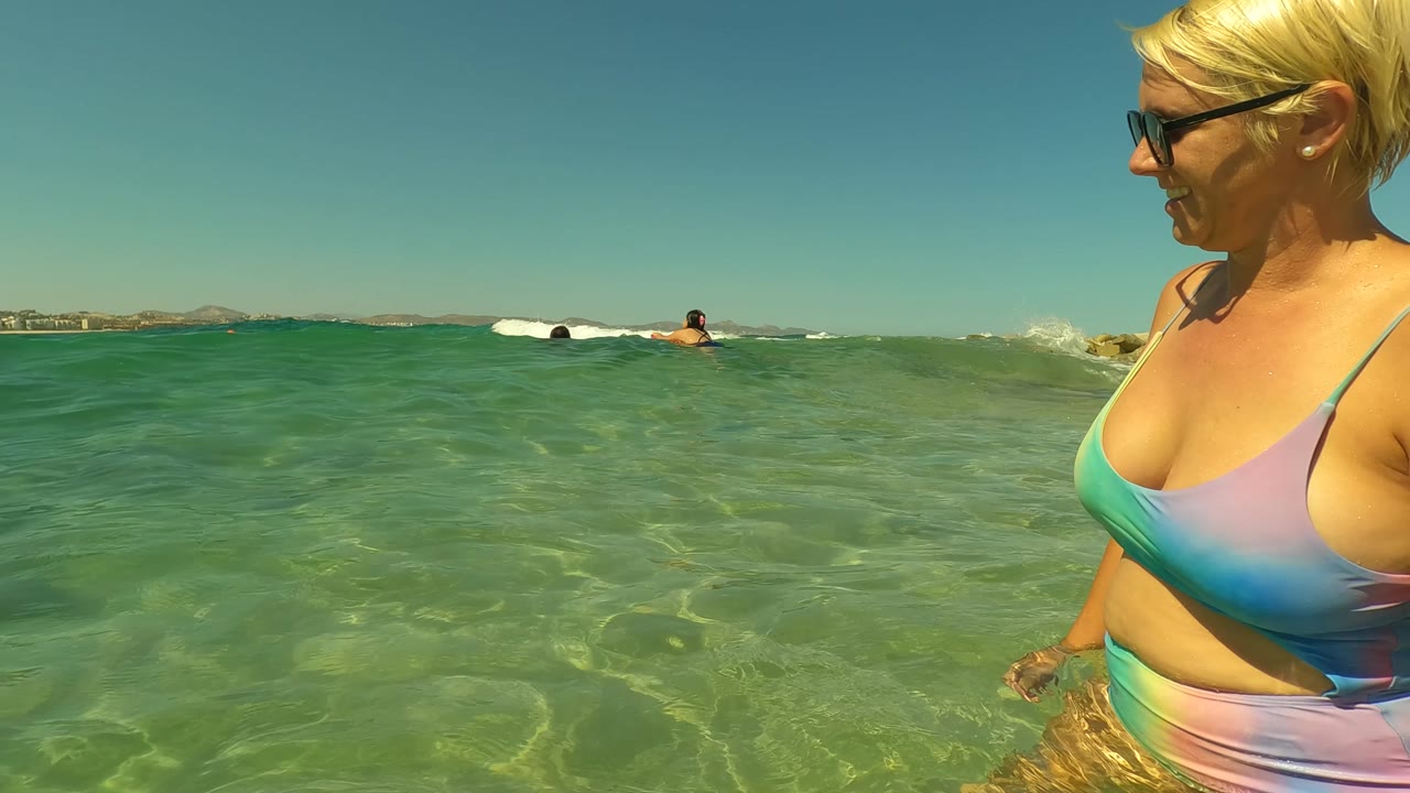 A woman wearing sunglasses stands at the right edge of the frame in clear shallow ocean water in San Jose del Cabo, Mexico, with swimmers and a low coastline visible across the horizon.