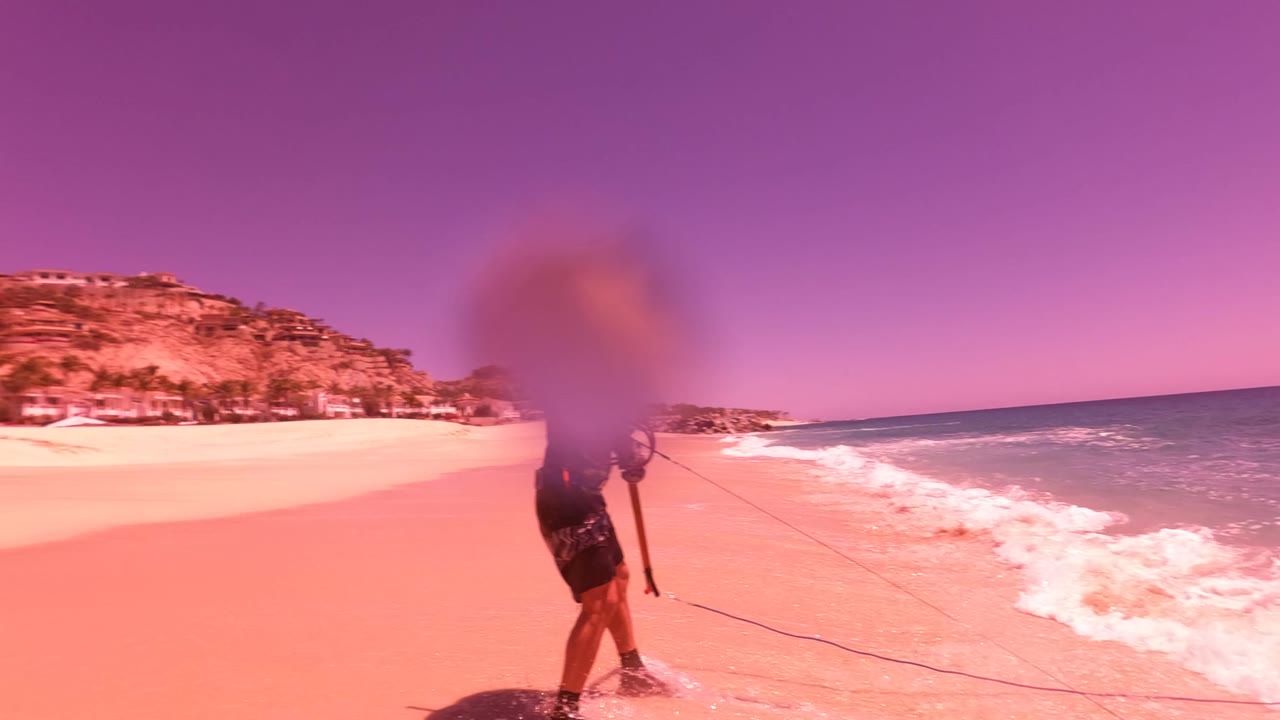 A person walks away on a sandy San Jose del Cabo beach beside breaking waves, with hillside resort buildings in the distance and a pink tint over the scene.