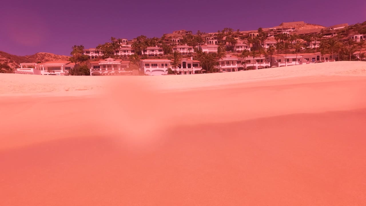 Empty pink-tinted beach sand spans the frame in San Jose del Cabo, with hillside resort buildings across the top and no visible people.