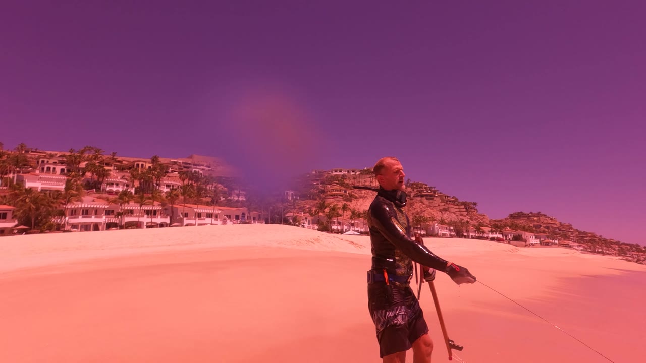 At 0:00 in San Jose Del Cabo BCS Mexico, a man in dark water gear holds a fishing rod on pink-tinted sand with coastal buildings and palms behind him.