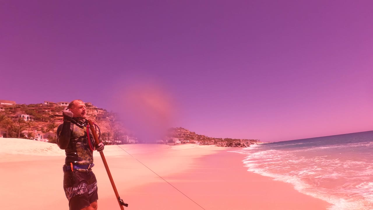 At 0:01 in San Jose Del Cabo BCS Mexico, the fisherman stands closer to center with ocean surf at right, hillside buildings behind, and a round lens water spot over the scene.