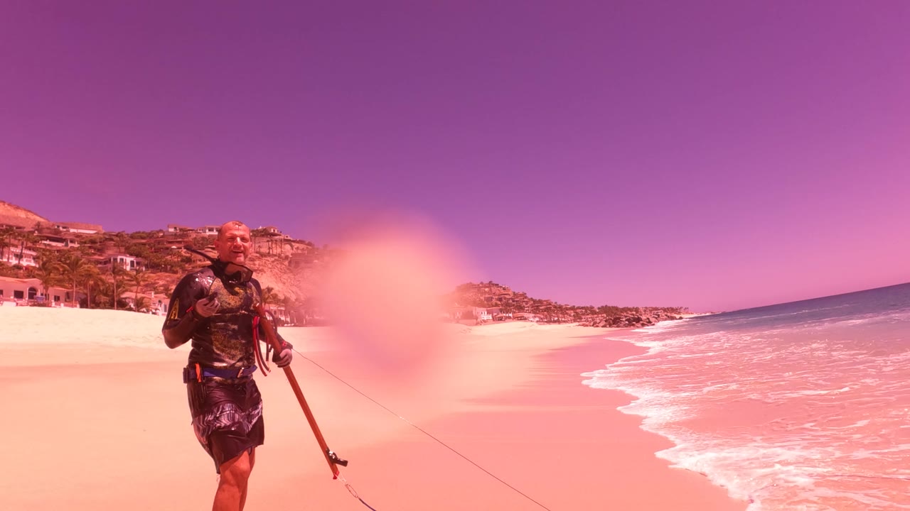 At 0:02 in San Jose Del Cabo BCS Mexico, the man with a fishing rod is framed near the surf as waves break along the right side and the beach curves past coastal buildings.