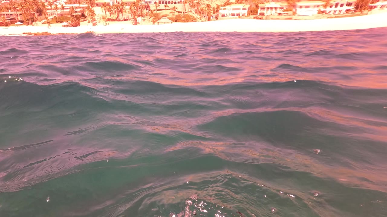 Pink reflections stretch across low ocean swell near San Jose del Cabo, Mexico, with a pale beach and distant coastal buildings visible above the horizon line, no visible people.