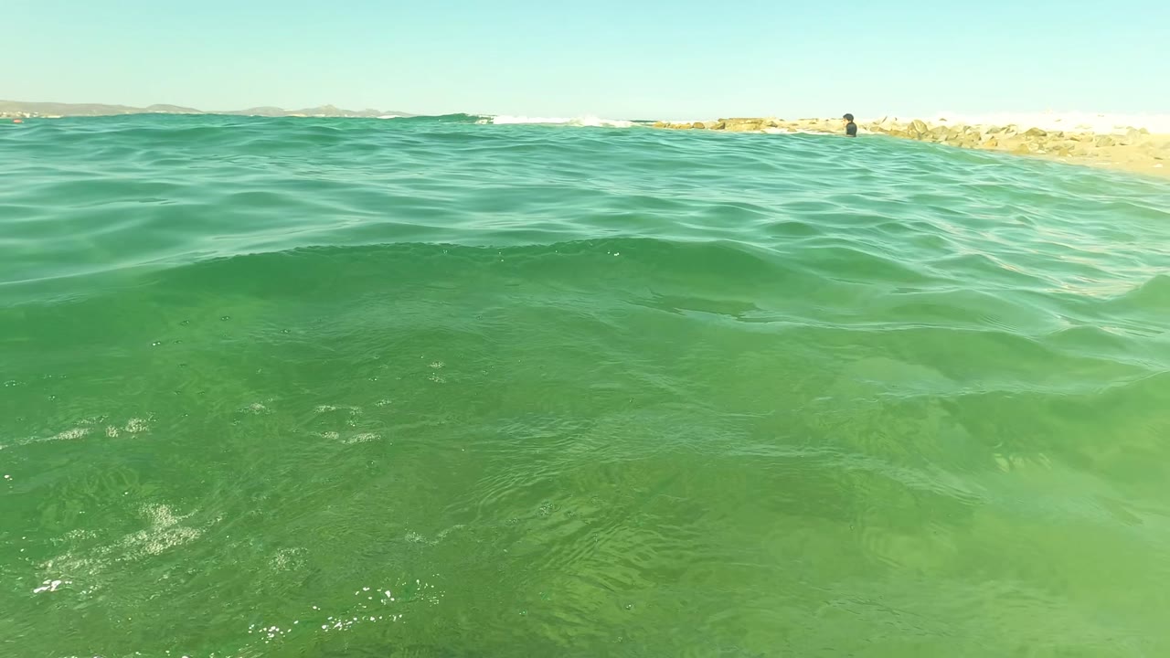 Water-level view of clear green ocean in San Jose del Cabo, Mexico, with small ripples, a low rocky breakwater, distant shore, and no visible people.