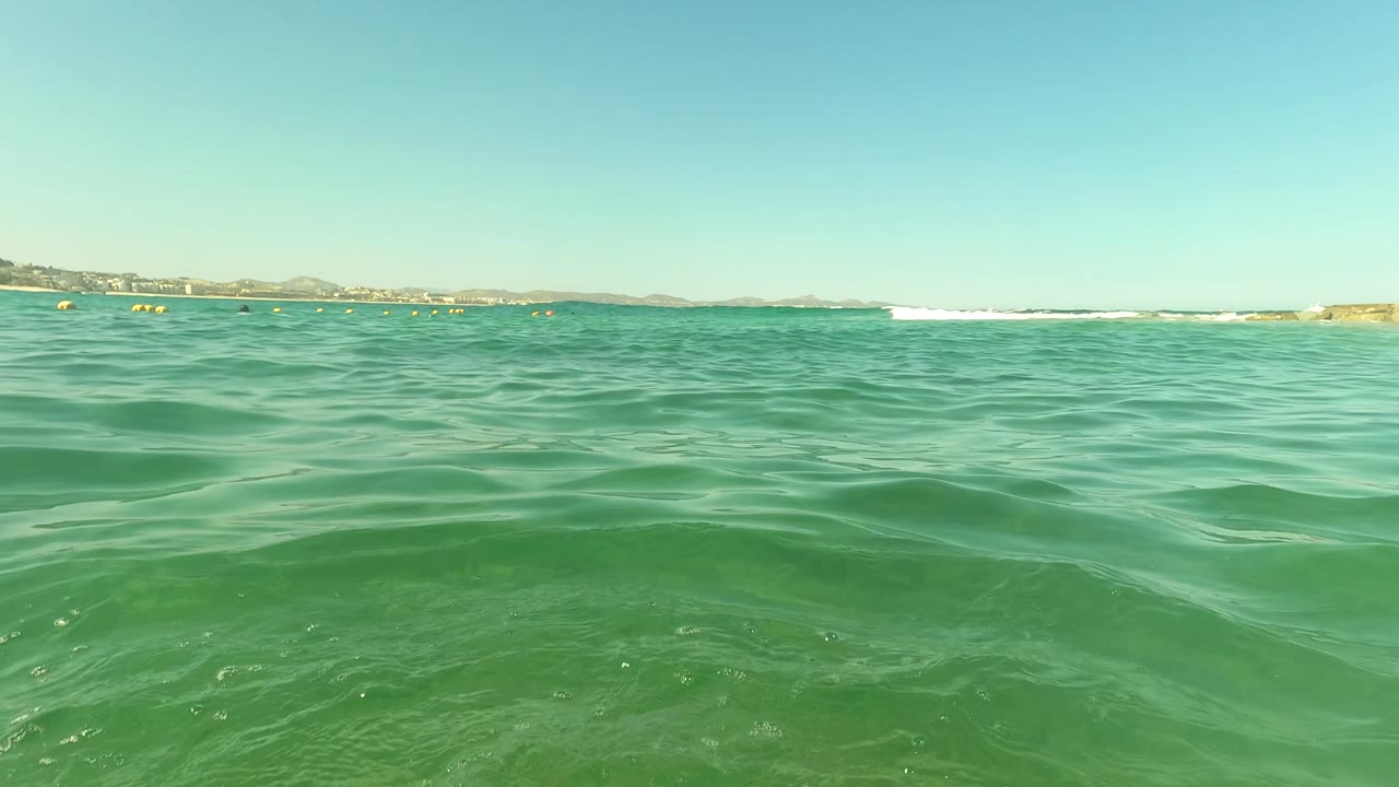 Water-level view in San Jose del Cabo, Mexico, with clear green waves, a distant shoreline, yellow floating markers, and no close visible people.