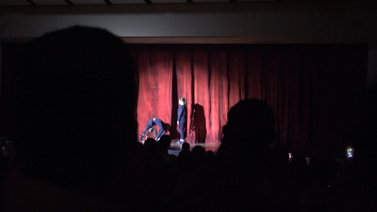 In San Jose Del Cabo BCS Mexico, two performers on a lit stage start an acrobatic sequence before a red curtain while the audience appears as dark silhouettes.