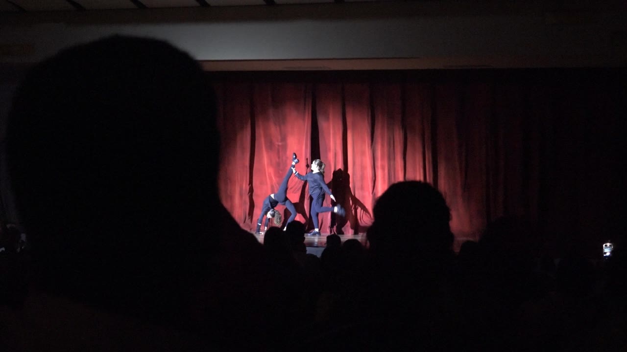 In San Jose Del Cabo BCS Mexico, two performers in dark clothing form a more extended acrobatic pose under stage light with the red curtain behind them.