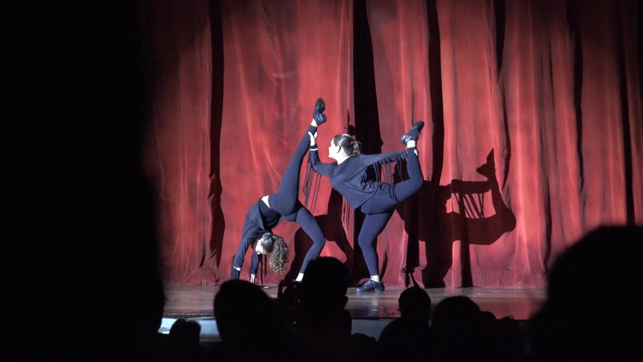 Two dancers in dark blue outfits perform a synchronized acrobatic pose on a red-curtain stage in San Jose del Cabo, Mexico, with audience silhouettes low in the foreground.