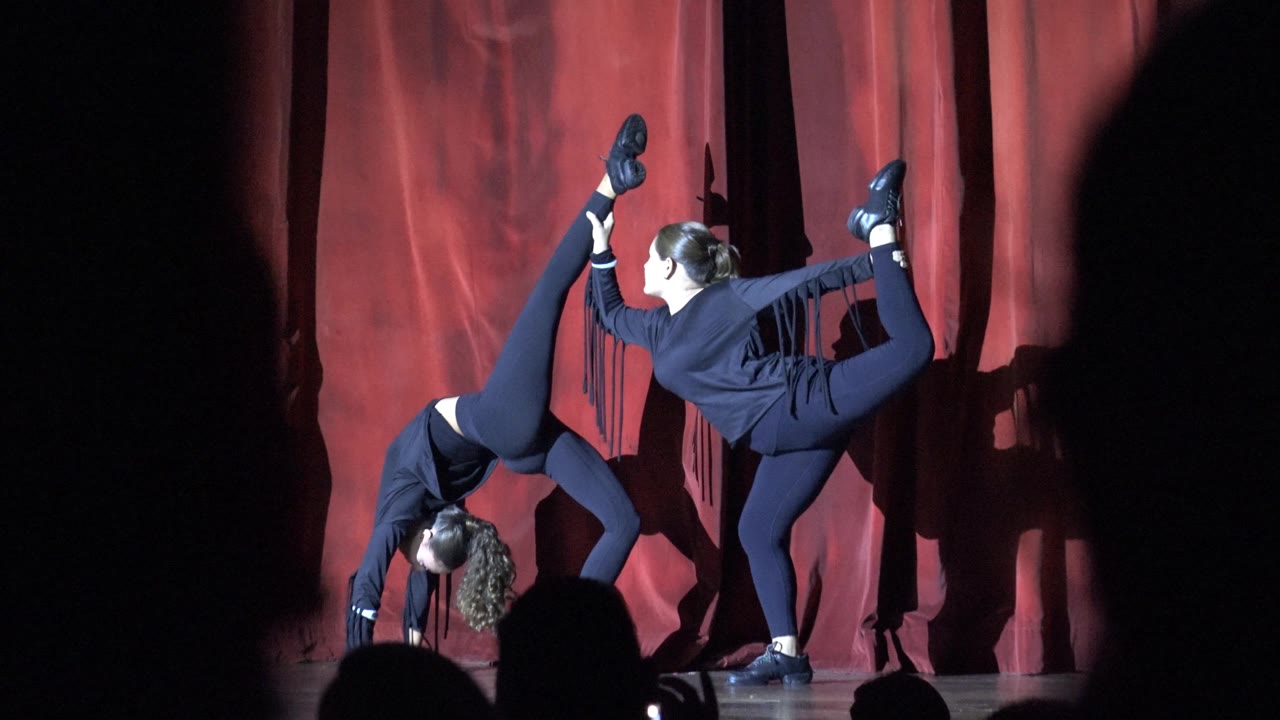 Two dancers in dark outfits perform an acrobatic pose on a red-curtain stage in San Jose Del Cabo BCS Mexico, with audience silhouettes across the lower foreground.