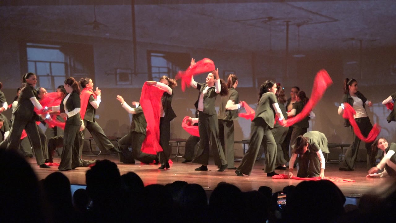 Wide frame from San Jose Del Cabo BCS Mexico showing dancers in green and white outfits spread across the stage while red scarves move above audience silhouettes.
