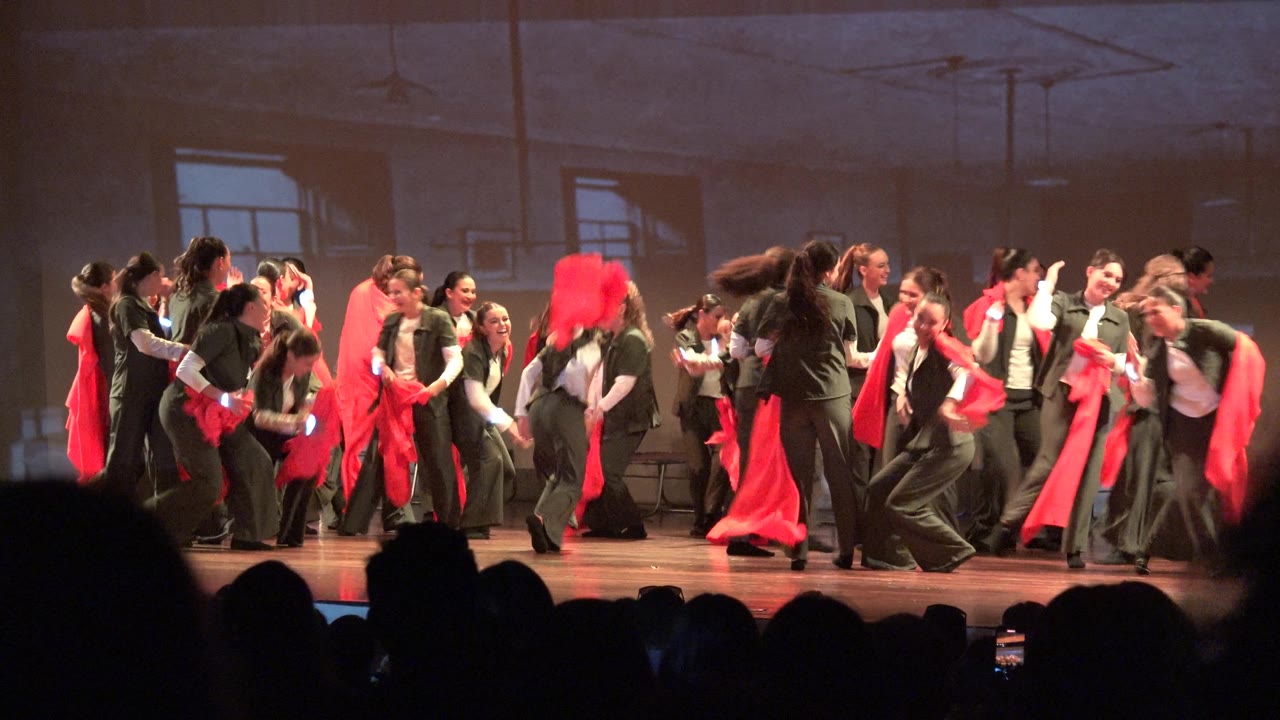 Wide theater view in San Jose Del Cabo BCS Mexico showing a large group of dancers in green outfits moving with red scarves while audience silhouettes line the foreground.