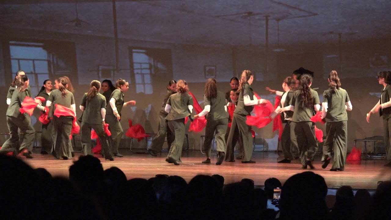 Dance group crosses the stage while holding red scarves.