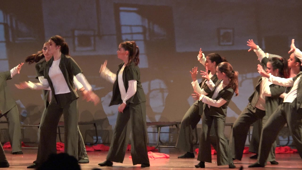Dancers on a lit stage in San Jose del Cabo BCS Mexico gesture outward in unison, with red fabric props on the floor and audience silhouettes below the frame.
