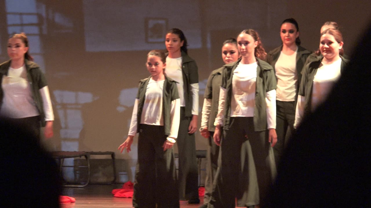 Dance group facing forward on stage with dark audience heads in front.
