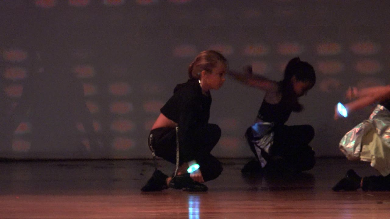 In San Jose Del Cabo BCS Mexico, the dancers begin to lift from their crouched poses, with one figure upright at the left edge and another arm movement blurred against the colored projection wall.