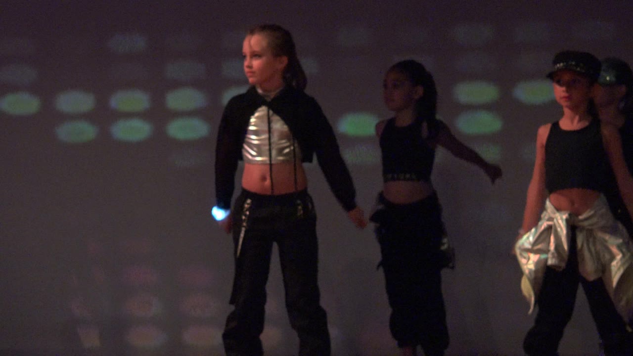 Children perform on a stage in San Jose Del Cabo BCS Mexico, with black outfits, silver clothing details, and soft colored light patterns on the backdrop.