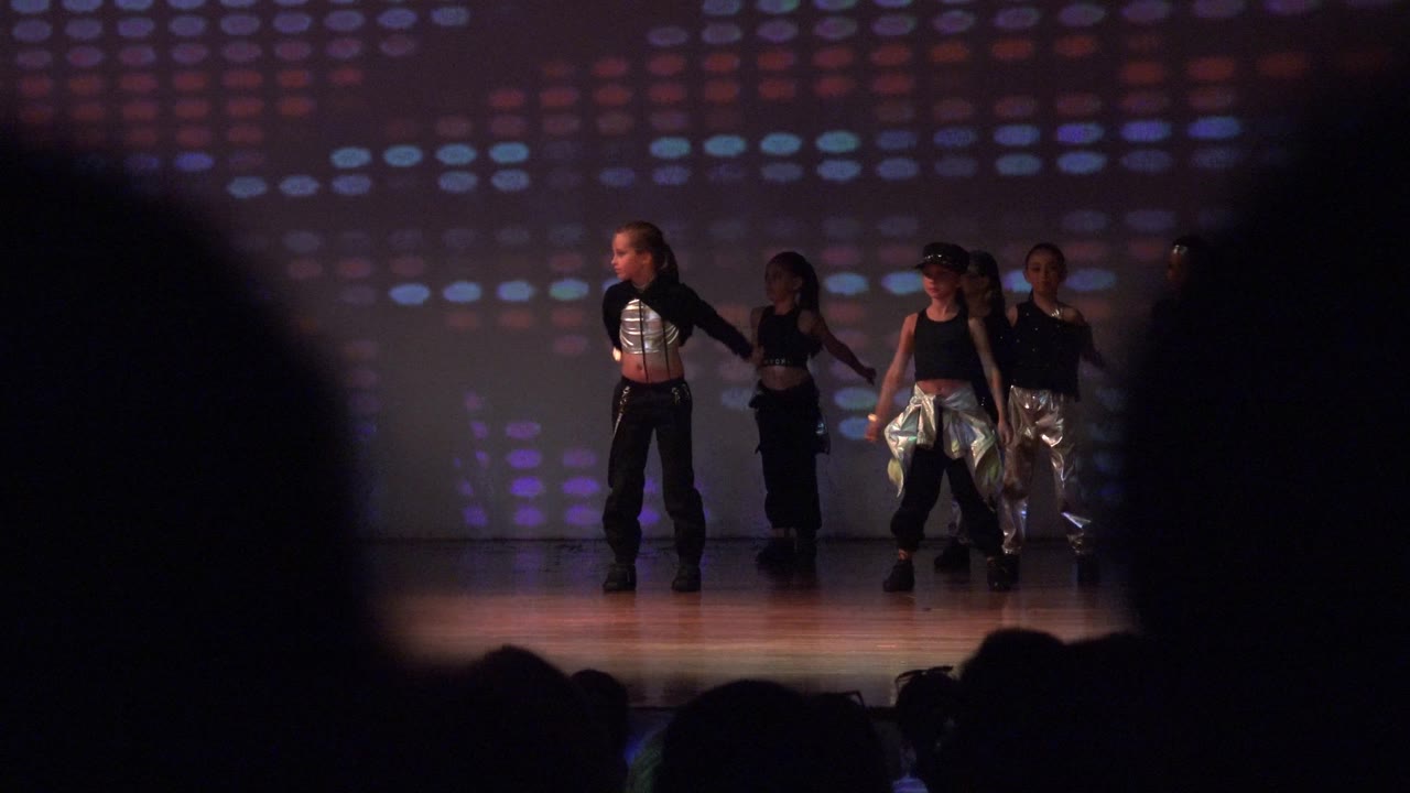 Wide stage view of children dancing behind audience silhouettes.
