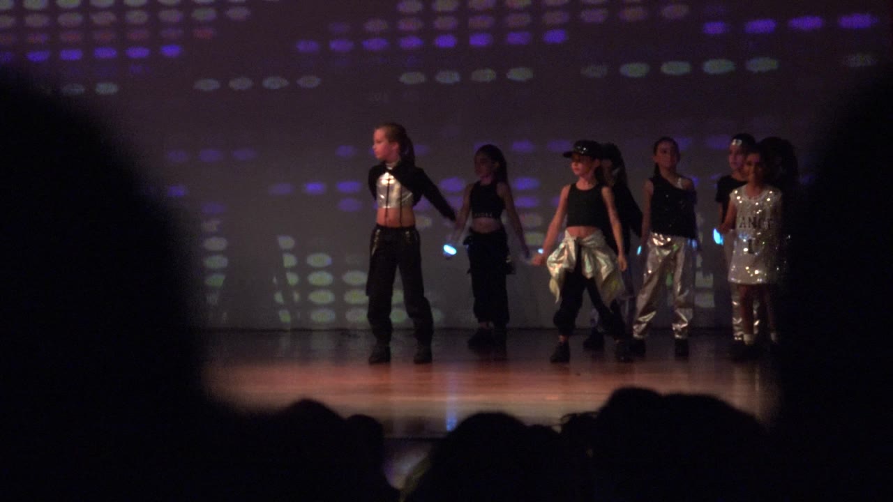 Several children stand across the stage in San Jose Del Cabo BCS Mexico, wearing black and silver performance outfits against a dotted colored light backdrop.