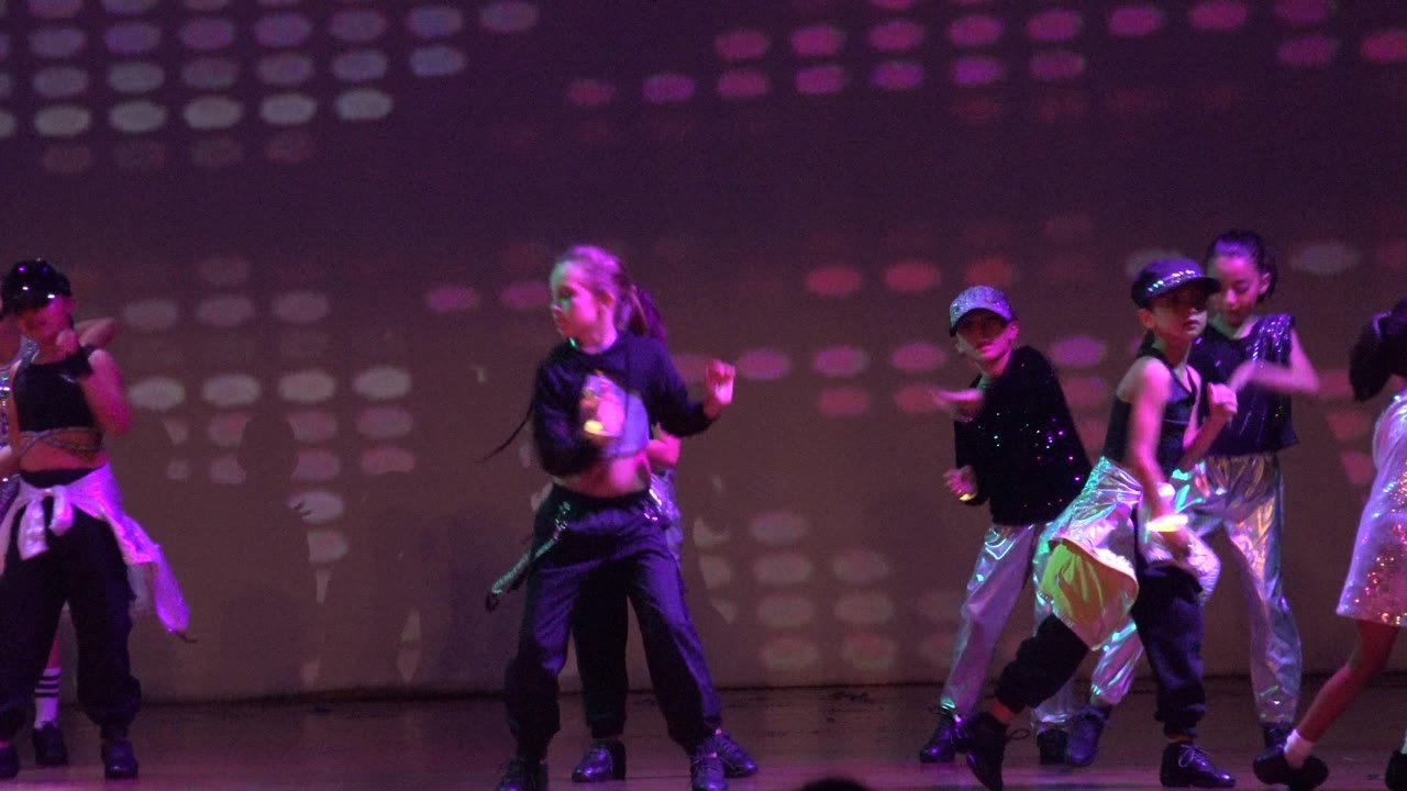 Children perform a group dance routine on a San Jose del Cabo BCS Mexico stage, with black outfits, shiny accents, and purple-green patterned lighting behind them.