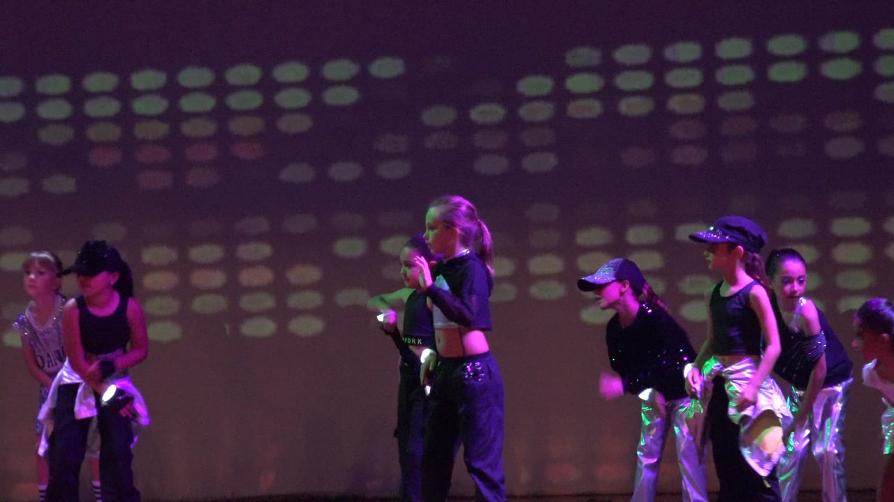 Dance performers on a dark stage in San Jose Del Cabo BCS Mexico, with several figures visible at the left and colored projection lights behind them.