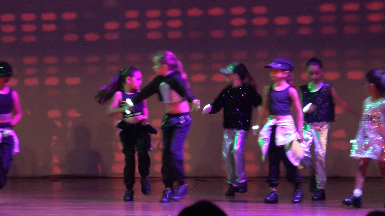Children in black tops, caps, silver pants, and a sparkly dress move across a stage in San Jose Del Cabo BCS Mexico with purple and green light on the backdrop.