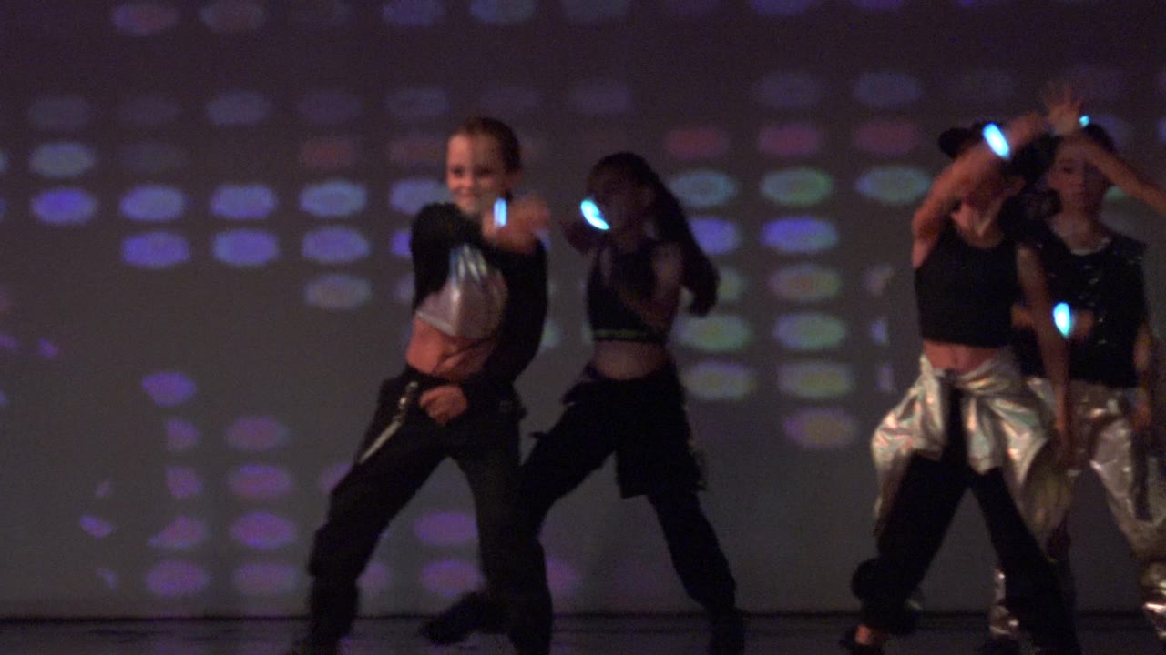 Three dancers perform under purple-blue light with glowing wristbands.