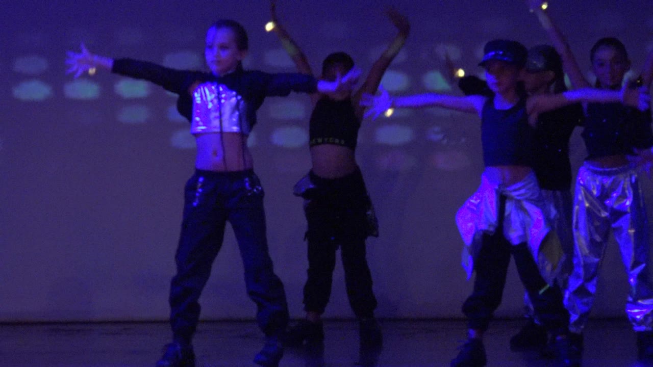 Teen dancers in San Jose Del Cabo BCS Mexico stand in formation under purple light, with one dancer extending an arm and colored projections on the rear wall.